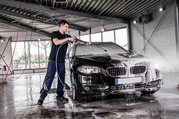 Schwarzes Auto in Waschstraße. Mitarbeiter wäshct das Auto mittels Hochdruckreiniger.